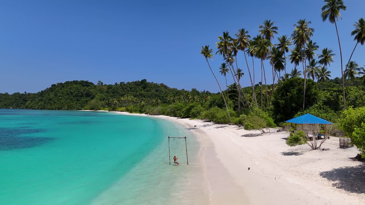 4k Drone video of a girl running to a swing on a tropical white sand beach, she sits on the swing and admires the beauty of Pompon Beach in the Banggai Islands, Sulawesi Indonesia