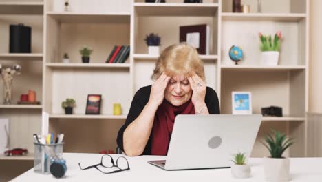 una mujer mayor y triste se sienta detrás de una computadora portátil, tratando de deshacerse de una migraña y dolor de cabeza masajeando sus sienes