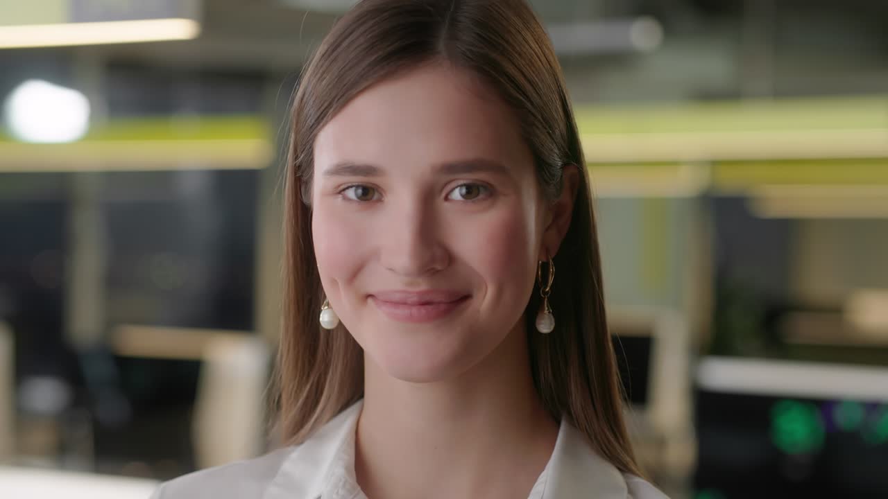 vista frontal de una hermosa mujer joven profesional con camisa blanca. retrato de una joven gerente de oficina que mira a la cámara y tiene buen estado de ánimo. fondo de oficina borroso