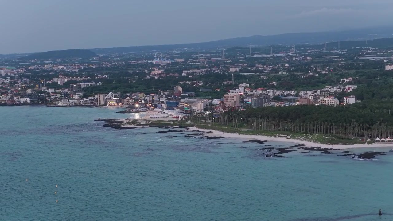Tomada cameo horizontal avançando para a esquerda ao longo da Praia de Hyeopjae, Ilha de Jeju, Coreia do Sul, mostrando a costa, o oceano, a praia, a vila e as rochas vulcânicas.