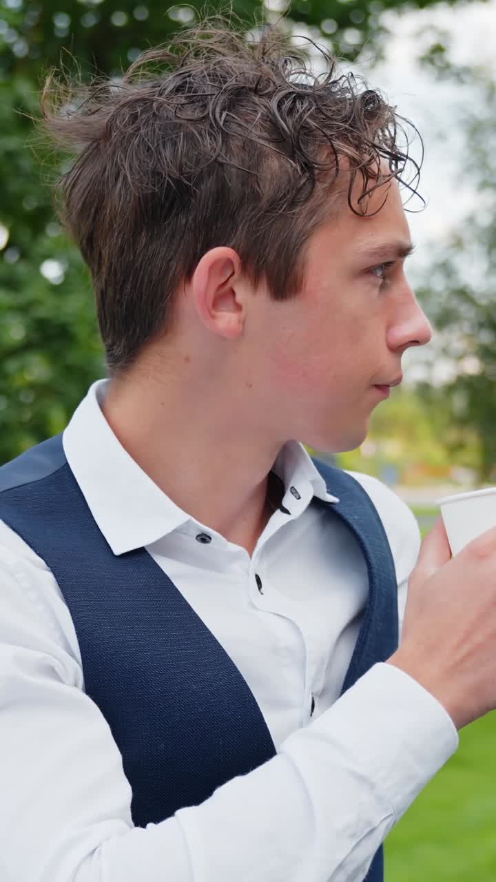 A vertical close-up shot shows a young man in formal business attire drinking coffee from a paper cup while standing outdoors