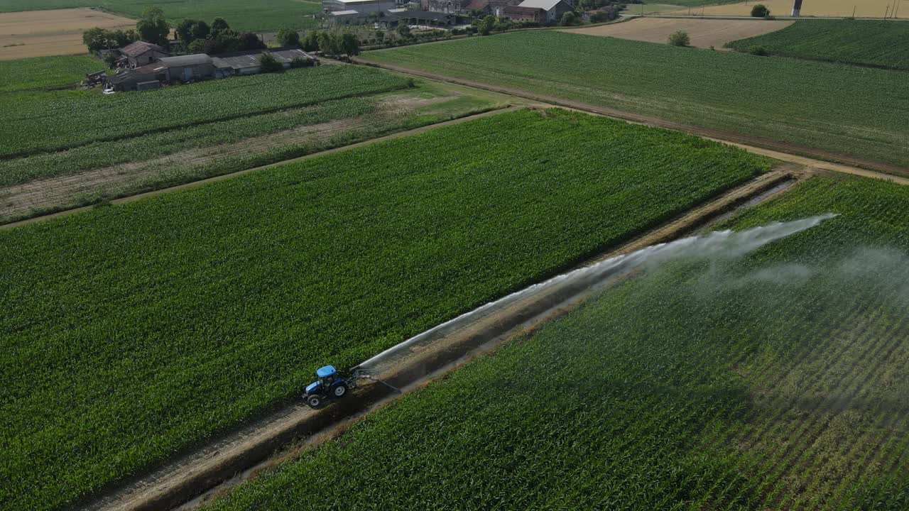 4K drone aerial rotating around a tractor in the foreground, gradually revealing a second tractor in the distant background irrigating farmland in Italy, with counterclockwise motion and water spray