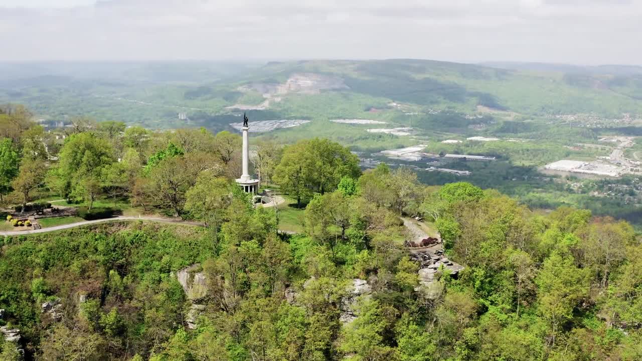 Drone capturing footage of solitary column standing tall amidst vast, lush green landscape during daytime in England, UK, surrounded by rolling hills, vibrant trees, and open fields