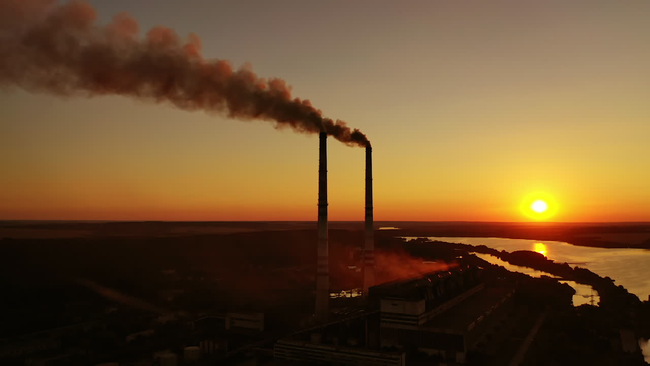 Air pollution. Dark smoke goes from industrial pipes at sunset. Silhouette of harmful factory near the river against the setting sun.