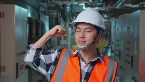 close up of asian male engineer with safety helmet flexing his bicep and smiling to camera while standing in engine control room, work of electrical generators