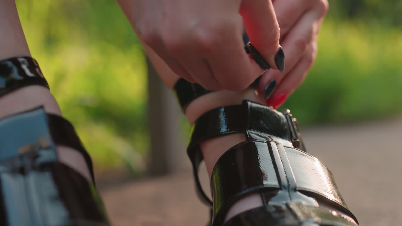 Sandalia con tira abrochada a mano con uñas pintadas en un banco iluminado por el sol, detalle de primer plano de la hebilla y el cuero brillante mientras el pie se prepara para caminar; estilismo íntimo y preparación capturados en la cálida luz de la tarde
