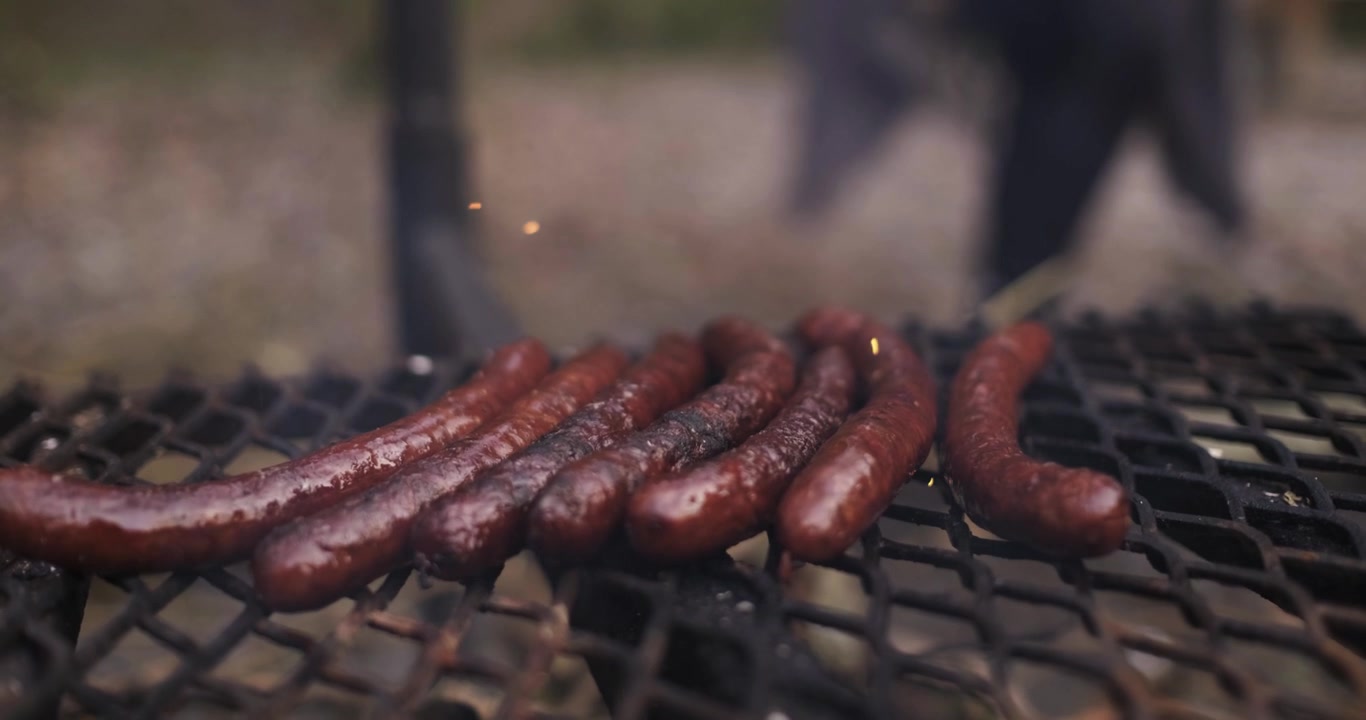 Close-up sosis yang sedang dimasak di atas api unggun di alam terbuka.