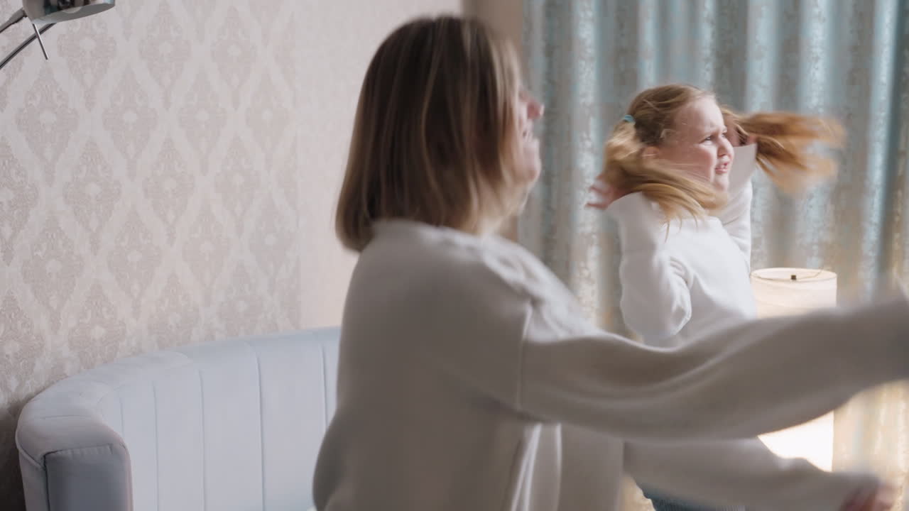 Mother and daughter in white sweaters share playful dance moment in cozy bedroom with soft lighting and patterned wallpaper, expressing joy, energy, family bonding, carefree lifestyle, connection indoors