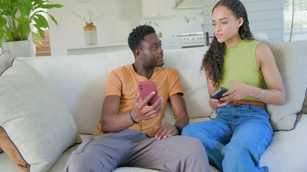 Casal afro-americano sentado no sofá, homem de camiseta laranja mostrando um telefone rosa, mulher de braços cruzados.