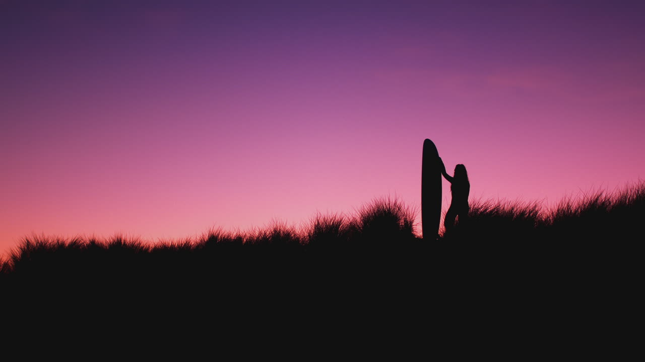 silueta de una mujer surfista de pie sosteniendo una tabla de surf en las dunas contra el sol poniente