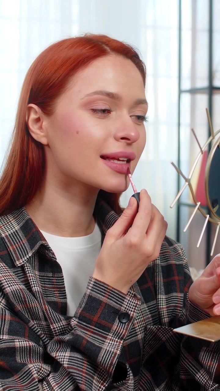 Young pretty woman applying makeup looking in mirror smiling while drawing lips with lipstick gently