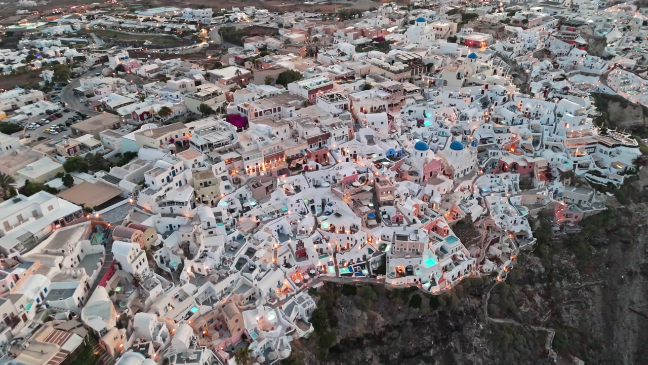 Un fitto gruppo di chiese dalle cupole blu di Oia, case rupestri e piscine luminose riempie il bordo della scogliera mentre la telecamera si allontana all'alba