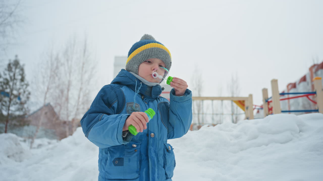 Giovane figlio nella neve che fa bolle durante un tranquillo gioco invernale, indossa un cappotto blu e un berretto di lana, tiene in mano una bacchetta verde per bolle e una bottiglia, cielo coperto e parco giochi sullo sfondo, espressione concentrata
