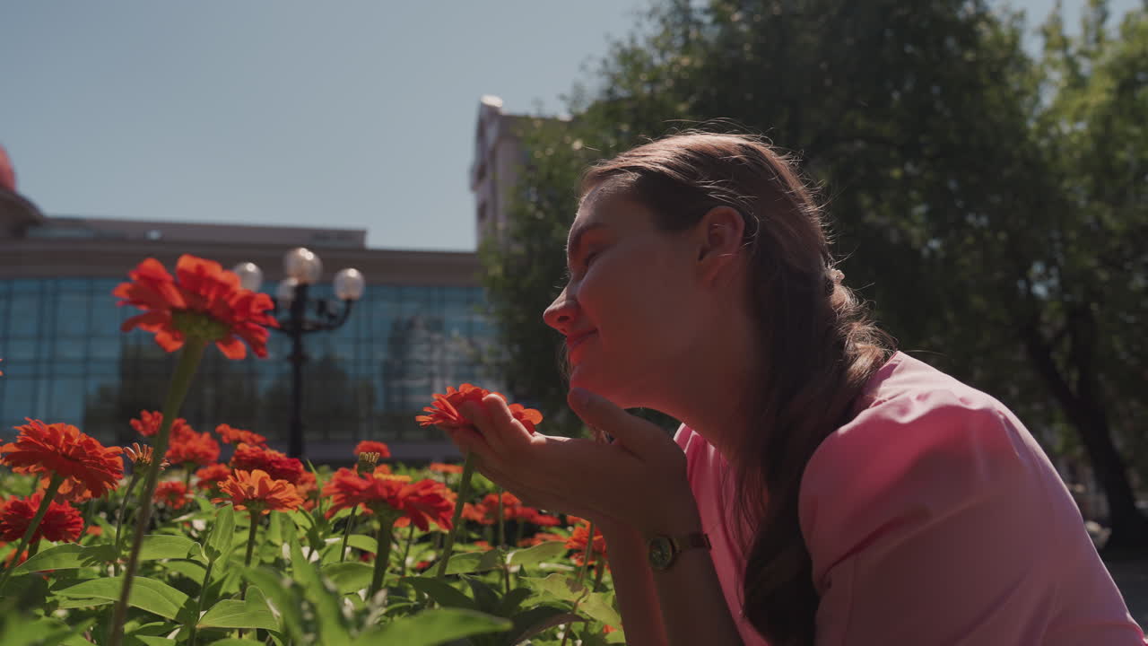 Une femme caucasienne dans un jardin urbain ensoleillé sentant des géraniums, robe rose, cheveux tressés, profil rétroéclairé, sourire doux, bâtiment de la ville et lampadaire en arrière-plan, fleurs rouge vif, ambiance estivale chaleureuse