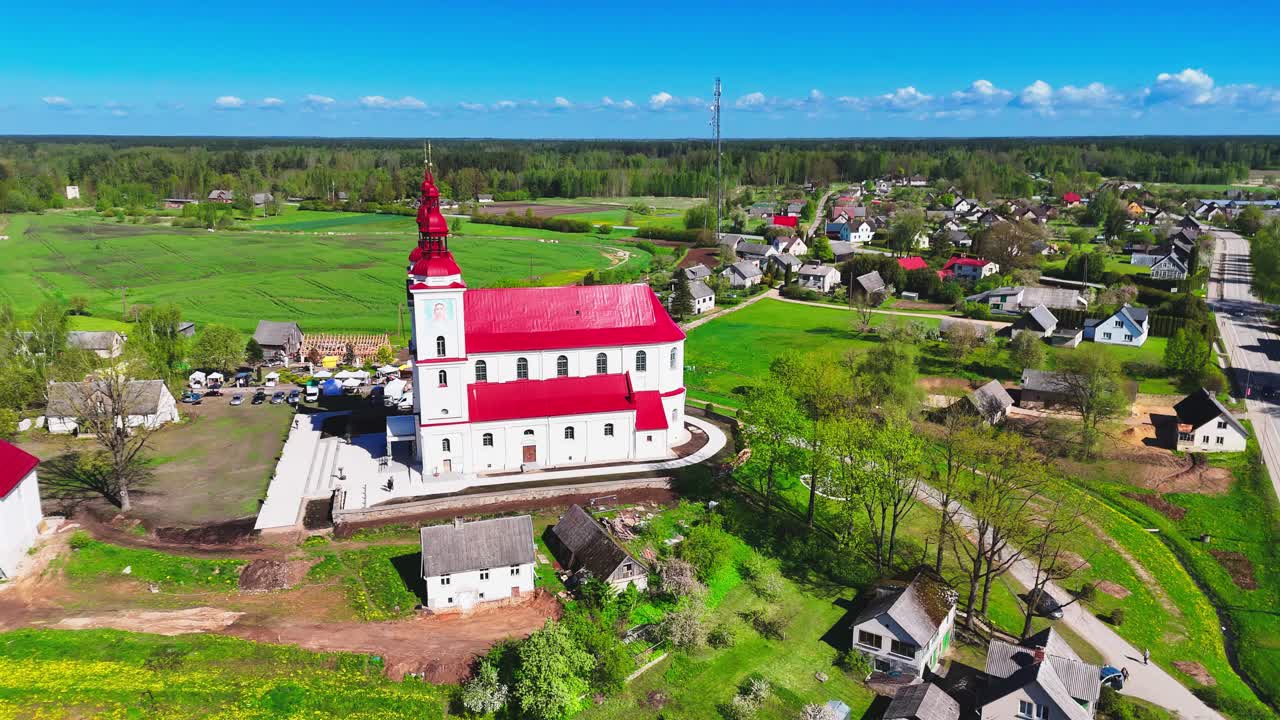 A historic white church with a vivid red roof rises above a peaceful rural village, with fields, cottages, and country roads stretching across the green European countryside.