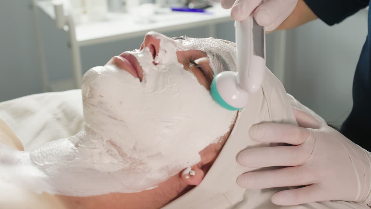 Wellness technician wearing protective gloves uses electric sculpt massaging tool to gently massage white mask on model face under bright spa lamp in modern clinic for relaxing skin therapy