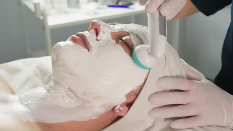 wellness technician wearing protective gloves uses electric sculpt massaging tool to gently massage white mask on model face under bright spa lamp in modern clinic for relaxing skin therapy