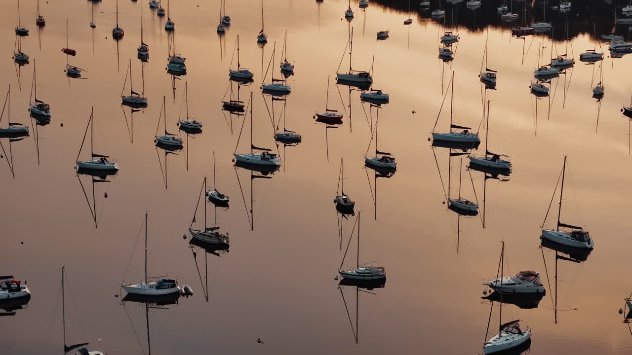 Sailboats on Windermere Lake with dazzling orange sky reflecting across serene water reflections of masts anchored on water.