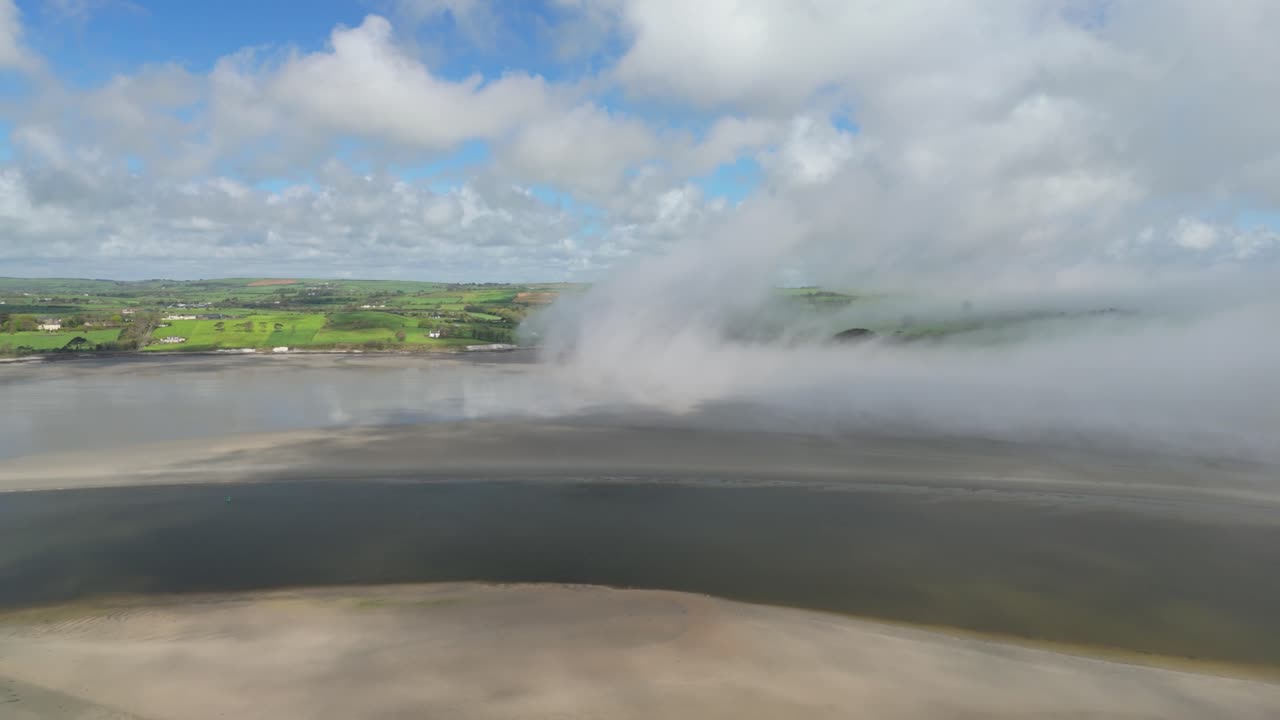 Morning ocean mist lifting from a sandy ocean bed on low tide in coastal Ireland with sunlit green hills, an aerial footage