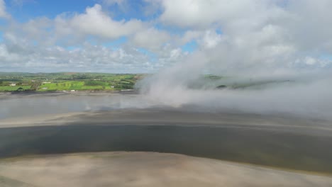morning ocean mist lifting from a sandy ocean bed on low tide in coastal ireland with sunlit green hills, an aerial footage