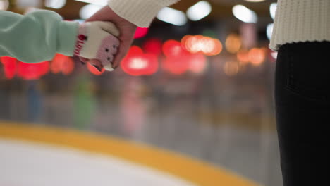 close-up view of a young girl holding her mom's hand while they skate together on an ice rink, the girl is wearing a mint green outfit, and the mom is dressed in a white sweater