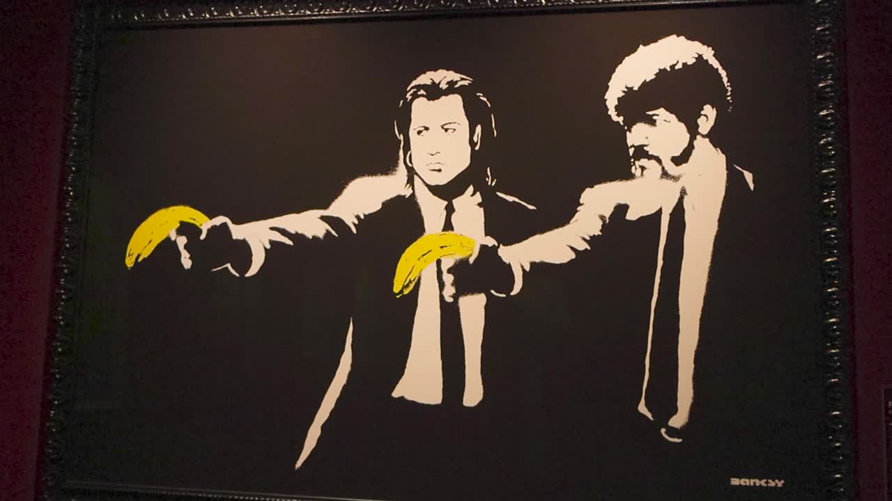 Banksy exhibition art showing two men from Pulp Fiction pointing yellow bananas as guns or weapons. Spray painting is black and white, while bananas are colorful, artwork is framed and on a wall.