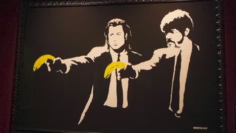 banksy exhibition art showing two men from pulp fiction pointing yellow bananas as guns or weapons. spray painting is black and white, while bananas are colorful, artwork is framed and on a wall.
