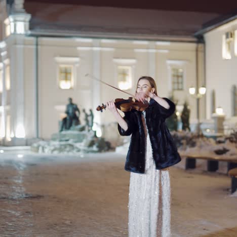 an attractive girl is standing in the square in a long white dress with sequins and playing the violin on the background of the monument and the buildings behind her in the evening. blurred background.