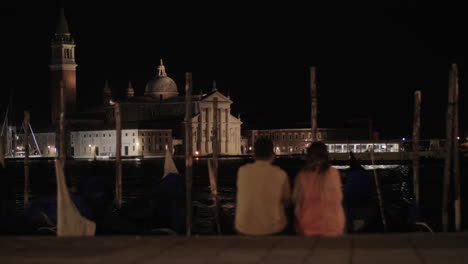 Una-Pareja-Sentada-En-El-Terraplén-Frente-A-La-Vista-Nocturna-De-Venecia