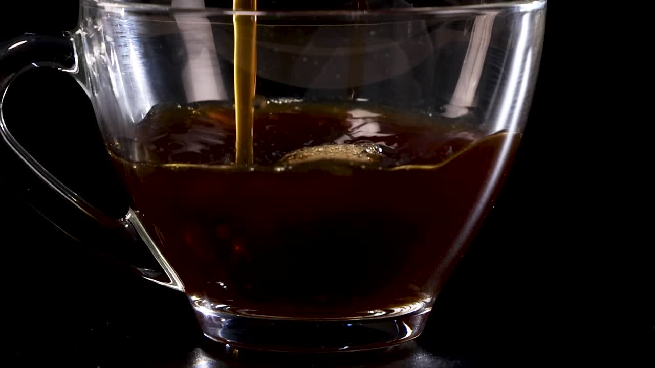 Un flusso costante di caffè nero riempie una tazza di vetro trasparente su uno sfondo nero