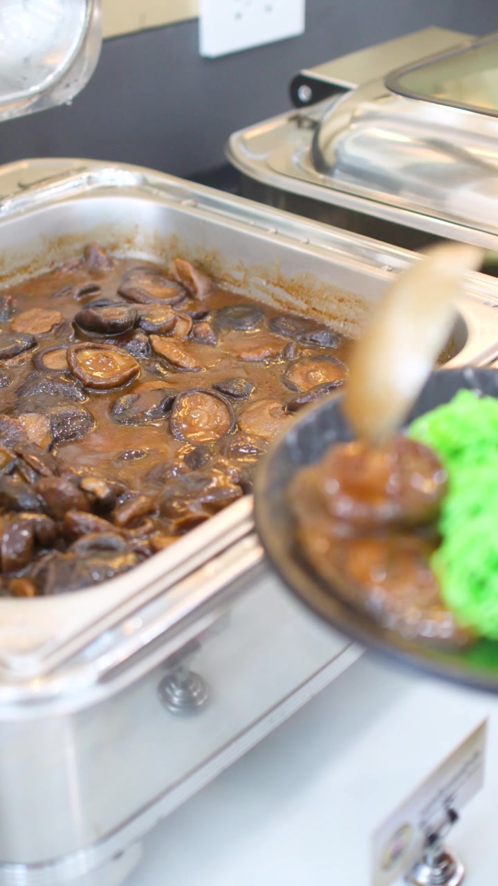 Un addetto al catering preleva funghi shiitake da uno scaldavivande in acciaio inossidabile e li serve su noodles verdi