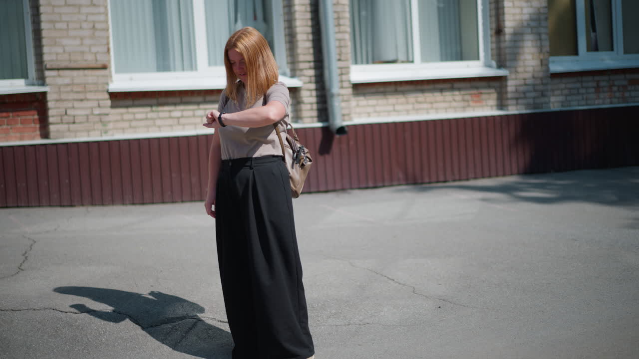 Young adult standing under bright summer sun near building, looking around and checking time with fitness bracelet, appearing tired and thoughtful, long shadow visible on sunny pavement outdoors