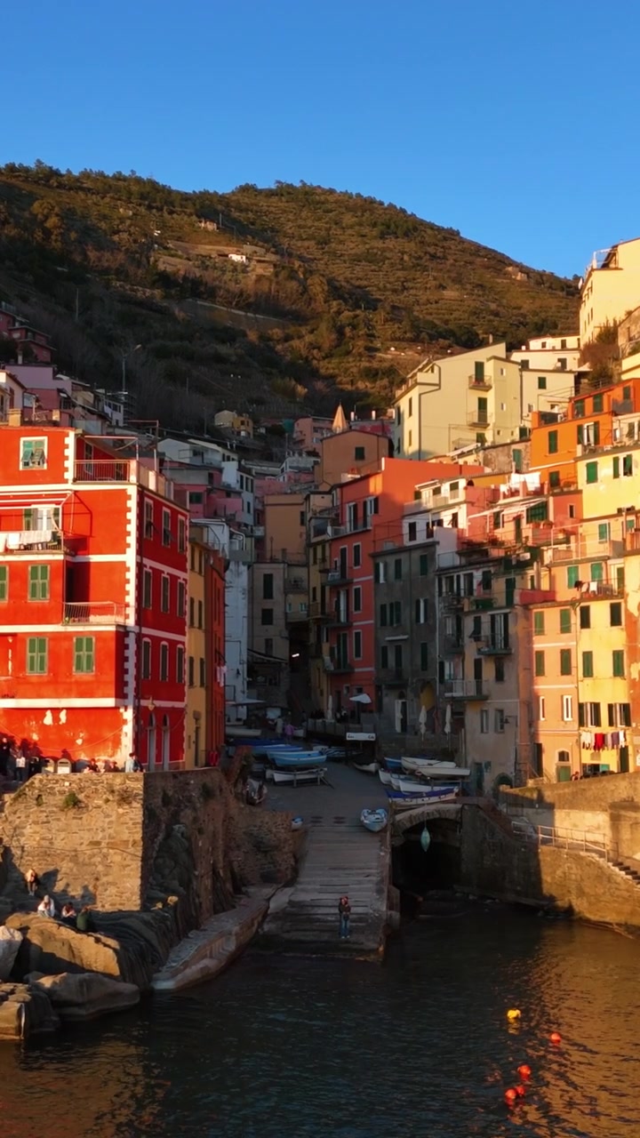 Bâtiments colorés à flanc de colline et petit port sous la lumière dorée du coucher de soleil, scène de village côtier.