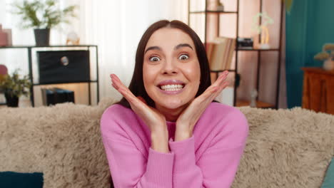 young woman sitting on sofa raising eyebrows opening mouth surprised amazed shocked by unexpected