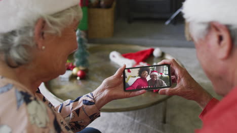 pareja caucásica mayor sonriente usando un teléfono inteligente para una videollamada de navidad con la familia en la pantalla