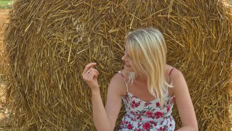 a young beautiful blonde woman sitting along hay rolls in a farm and playing with the same enjoying the wonderful nature