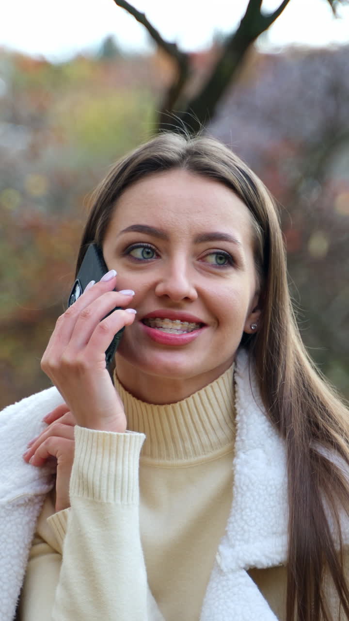 Mid-age Caucasian woman with dark hair speaks on the phone. Positive lady having phone conversation in the autumn park. Blurred backdrop. Vertical video