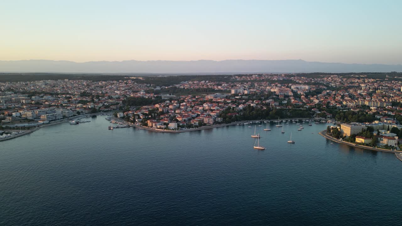 크로아티아의 자다르 시의 해안선을 따라 푸른 아드리아 해, 세일보트, 마리나, 주택 및 건물 및 해가 지는 먼 산 벨리 ⁇ 과 함께 항공 파노라마