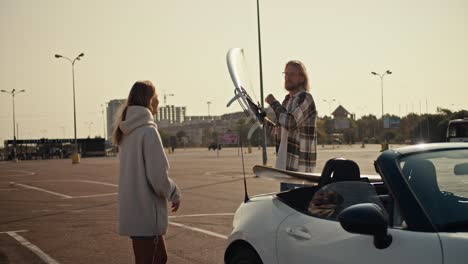 Ein-Blonder-Typ-In-Einem-Karierten-Hemd-Und-Mit-Brille-Gibt-Seiner-Blonden-Freundin-In-Einem-Weißen-Sweatshirt-Ein-Weißes-Surfbrett-Aus-Seinem-Weißen-Cabrio-Und-Nimmt-Seins-Mit-Auf-Den-Großen-Platz-In-Der-Stadt