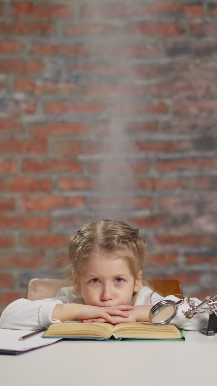 Upset little girl with smoke rising above head tired of hard home task reads textbook putting chin on hands near laptop at table in light room