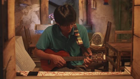 Looking-through-an-open-window-at-a-young-Asian-male-playing-a-ukulele-while-on-a-chair-in-a-rustic-room