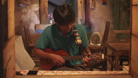 looking through an open window at a young asian male playing a ukulele while on a chair in a rustic room