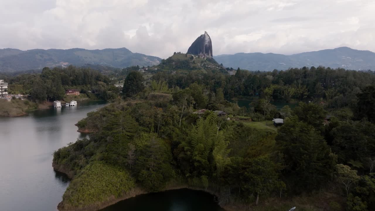 Drone shot of El Peñón de Guatapé (La Piedra Rock) with lakes and mountains on a cloudy day with helicopter flying across rock