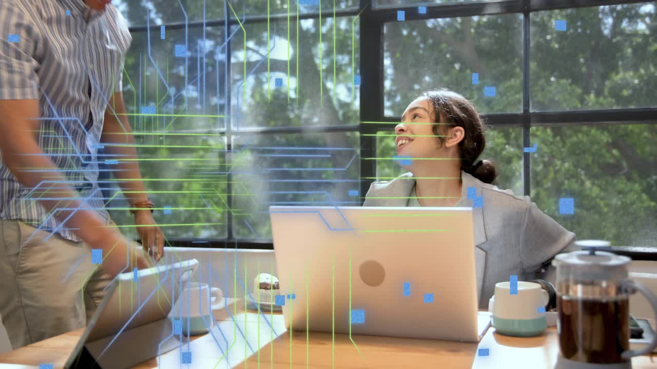 Woman typing at laptop while peer approaching, speaking, animated data overlays aiding tech work