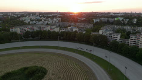 Corredores-De-Arnés-En-La-Pista-De-Carreras-De-Caballos-De-Tierra-En-Berlín,-Alemania-Al-Atardecer,-Toma-De-Seguimiento-Aéreo-Desde-Arriba