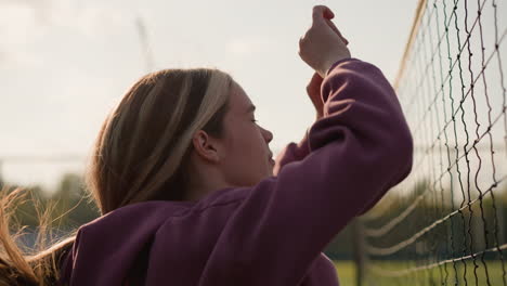 side view of lady in maroon hoodie training in front of volleyball net, sunlight reflecting off her face and hair fluttering with wind, background features trees and sports facilities