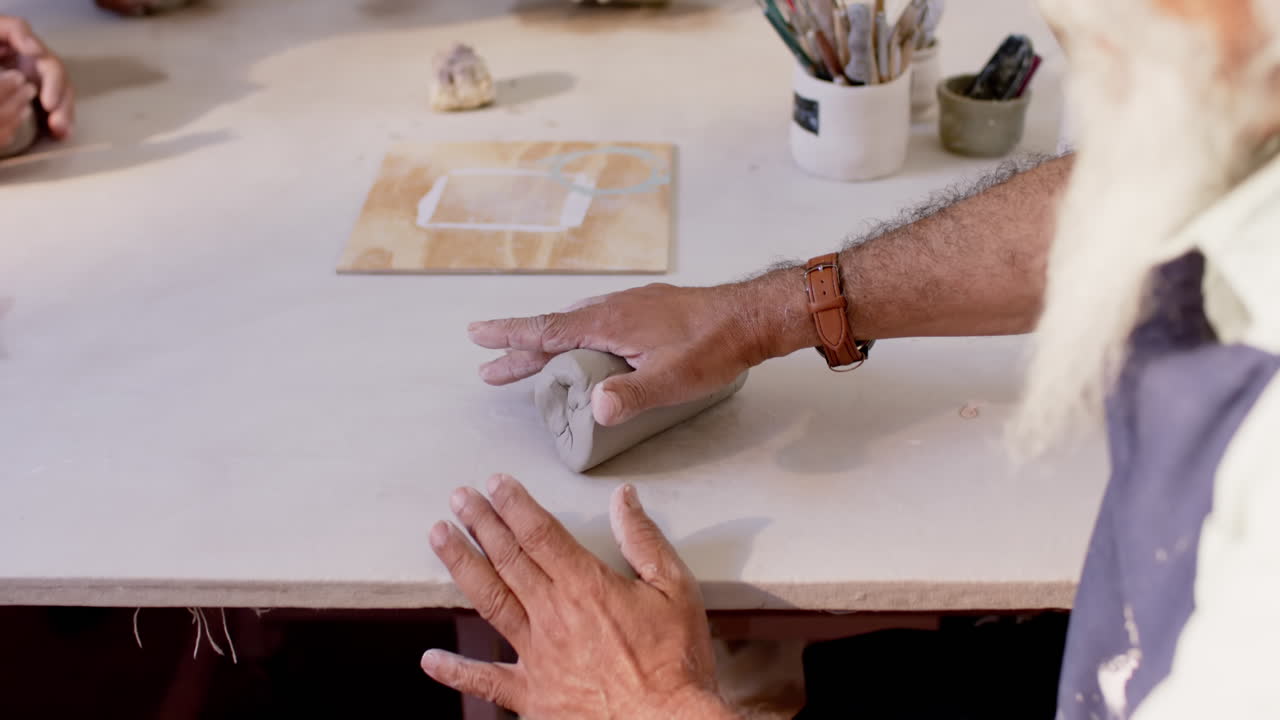 ceramista biracial enfocado con barba larga moldeando arcilla con las manos en el estudio de cerámica, cámara lenta