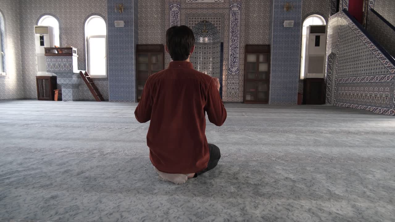 Back view of a young bearded man sitting inside mosque praying to god with open hands, Muslims who go to historical and holy places pray