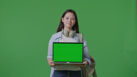 asian teen girl student with a backpack smiling and showing green screen laptop to camera while standing in the green screen background studio