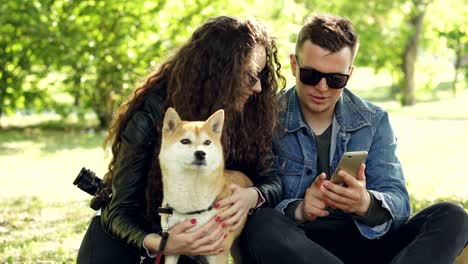 young woman is petting her dog sitting on grass while her boyfriend is showing her smartphone screen, people are talking and laughing. weekend in the park concept.